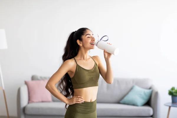 Hübsche Inderin in Sportkleidung trinkt zu Hause Wasser oder Proteinshake aus der Flasche — Stockfoto