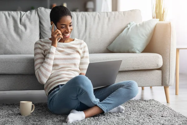 Vreugdevolle Afrikaanse Amerikaanse vrouw praten op mobiele telefoon en het gebruik van laptop thuis — Stockfoto