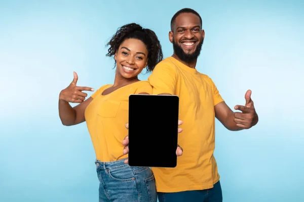 Alegre pareja negra sosteniendo la tableta, apuntando a la pantalla vacía con espacio para maqueta, la promoción de la nueva aplicación, sitio web —  Fotos de Stock