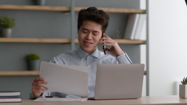 Mladý pozitivní asijský muž office manager při pohledu na dokumenty a mluvit s obchodními partnery prostřednictvím mobilního telefonu — Stock video