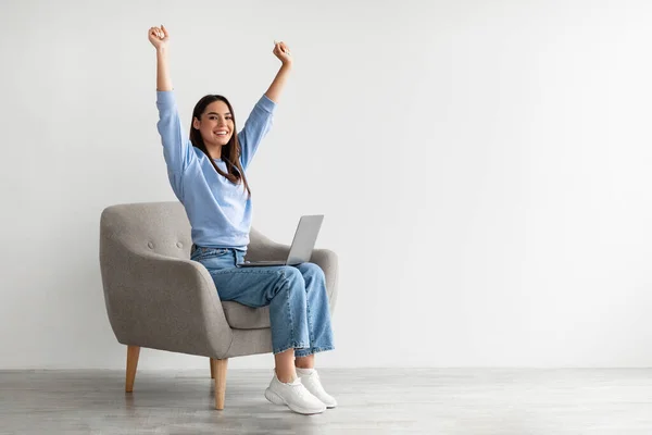 Mujer extática sentada en sillón con portátil, levantando las manos, haciendo un gesto SI, celebrando victoria o éxito en línea — Foto de Stock