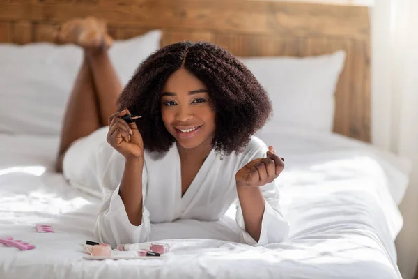 Wanita kulit hitam yang gembira berbaring di tempat tidur dengan jubah, mengoleskan cat kuku, membuat manikur, tersenyum di depan kamera — Stok Foto