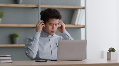 Genç Asyalı adam kulaklığını çıkarıyor ve düğüm köprüsünü ovuyor, müşterilerden bıkıyor ve rahatsız oluyor, internette çalışıyor.