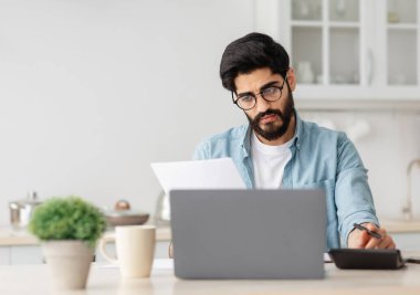 Stresli Arap adam ev bütçesini hesaplıyor, mutfakta oturuyor, genç adam mali kriz yaşıyor, borçları var.