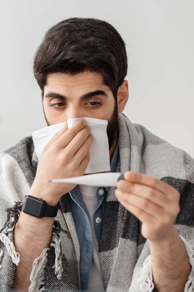 Seasonal infectious disease. Sick arab guy sneezing, measuring body temperature, sitting at home interior, crop, closeup — Stock Photo, Image