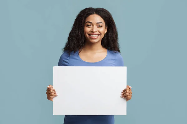 Lugar para o seu anúncio. Mulher negra alegre segurando cartaz em branco com espaço livre para propaganda sobre fundo azul — Fotografia de Stock