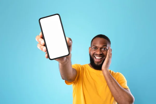 Homem negro animado apresentando smartphone com tela vazia, recomendando site ou anúncio, fundo azul, espaço para mockup — Fotografia de Stock