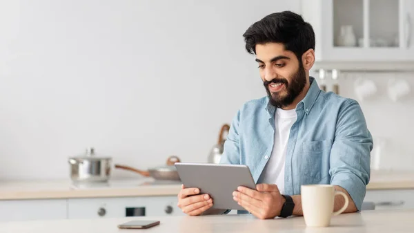 Veselý arabský muž pomocí digitální tablet při pití kávy v kuchyni doma a kontrolu sociálních sítí, kopírovat prostor — Stock fotografie