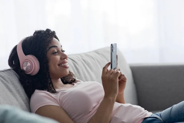 Lieto giovane signora nera in cuffie wireless digitando sul telefono in salotto interno — Foto Stock