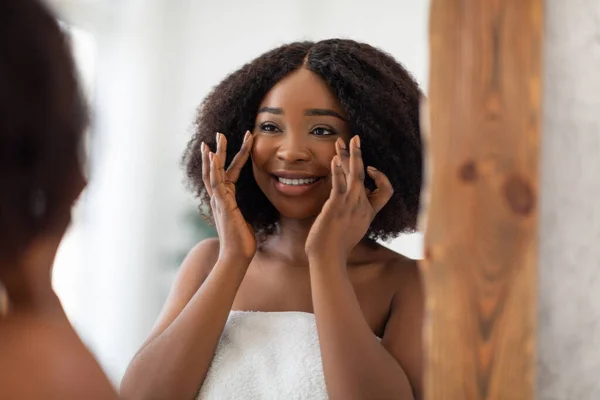 Charmante dame noire qui se sourit dans le miroir, vérifiant les premières rides, admirant la peau douce après la crème visage à la maison — Photo