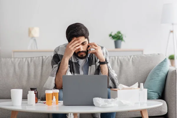 Sick millennial arab man covered in blanket sitting on couch, having conversation on phone while working on laptop — Stock Photo, Image