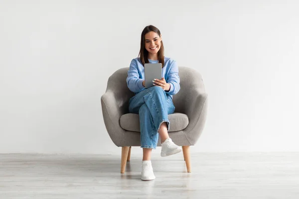 笑顔の若い女性の肖像画デジタルタブレットアームチェアに座って、白いスタジオの壁に対してオンラインで作業 — ストック写真