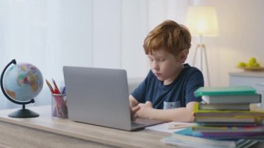 Evde eğitim kavramı. Dizüstü bilgisayarda daktilo eden sevimli küçük bir çocuk, evde kitaplarla ve küreyle oturuyor.