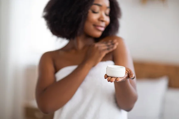 Donna nera femminile che tiene il vaso di crema per il corpo o il viso, applicando il prodotto per la cura della pelle dopo la doccia a casa — Foto Stock