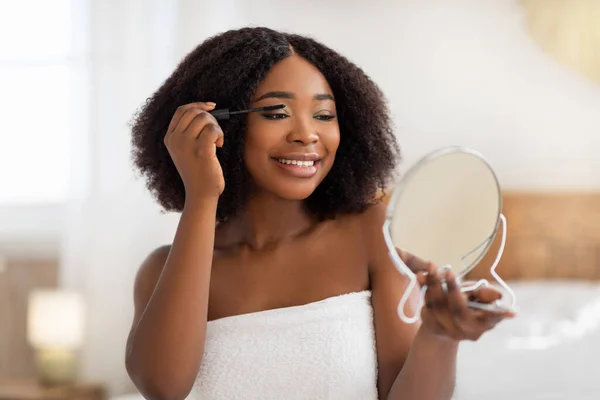 Skönhetssmink. Porträtt av glad ung afro kvinna tillämpa mascara efter bad eller dusch, titta i spegeln inomhus — Stockfoto