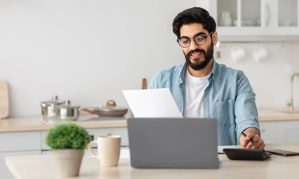 Portrait d'un homme arabe heureux lisant des documents d'assurance à la maison, assis à table dans la cuisine et comptant le budget de la maison — Photo