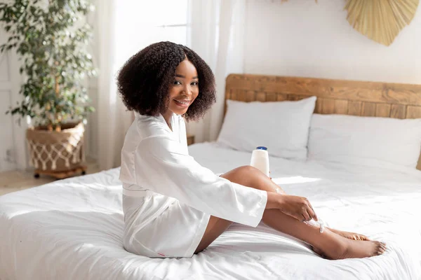 Wanita kulit hitam yang bahagia dengan jubah putih mencukur kaki dengan pisau cukur, menggunakan busa, duduk di tempat tidur, melakukan prosedur perawatan kulit dalam negeri — Stok Foto