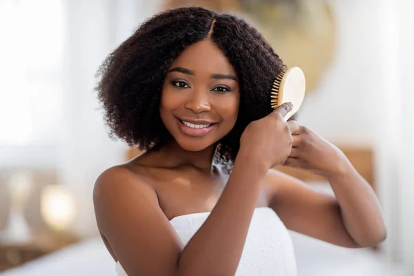 Retrato de bela jovem afro senhora escovando seu cabelo escuro, sorrindo para a câmera dentro de casa — Fotografia de Stock