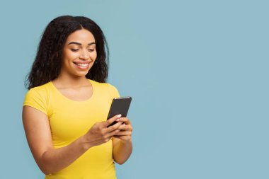 Mutlu Afro-Amerikalı kadın akıllı telefon mesajları ve iletişim ağlarını kullanarak mavi stüdyonun arka planında duruyor.