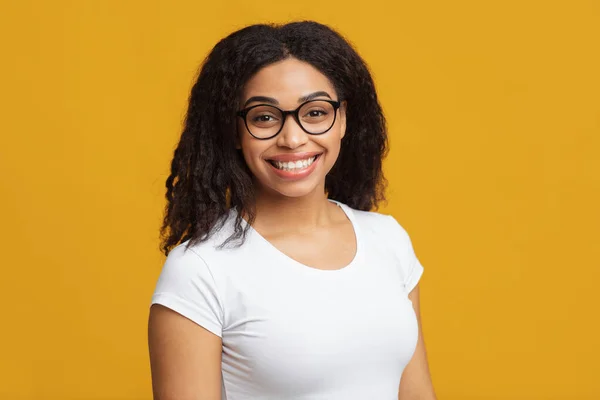 Sıradan biri. Gözlüklü siyah kız öğrencinin portresi kameraya bakıyor ve gülümsüyor, sarı arka plan — Stok fotoğraf