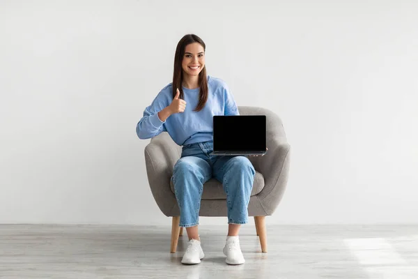 Donna caucasica che mostra computer portatile con schermo bianco, gesticolando pollice in su, seduto in poltrona contro muro bianco, mockup — Foto Stock