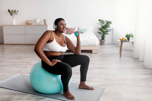 Mujer Afro con sobrepeso haciendo ejercicio con pesas, sentada en el fitball, escuchando música en auriculares en interiores —  Fotos de Stock