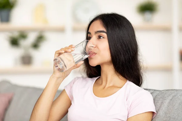 Blijf gehydrateerd. Aantrekkelijke jonge Indiase vrouw drinkwater uit glas op de bank in de woonkamer — Stockfoto