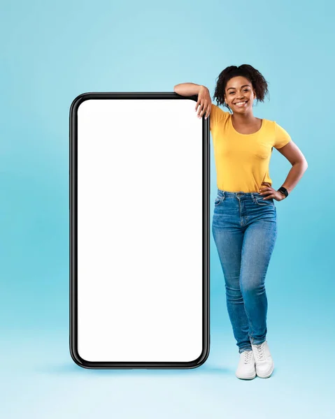 Büyük, beyaz ekranlı akıllı telefona yaslanan mutlu siyah kadın mavi arka plandaki kameraya gülümsüyor. — Stok fotoğraf