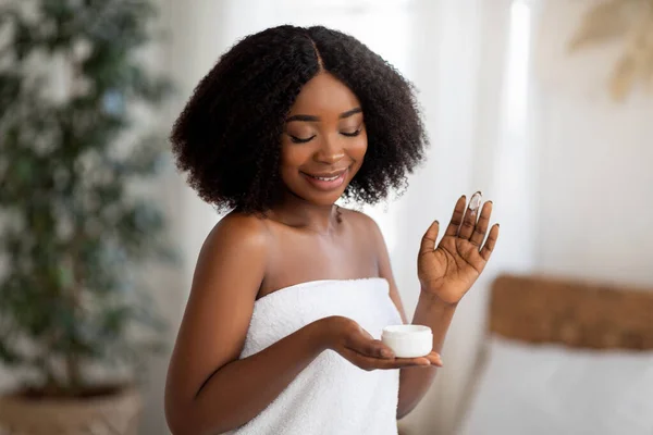 Νεαρή Αφρο-Αμερικανίδα ποζάρει με ένα βάζο ενυδατικής κρέμας, χαμογελαστή, φορώντας πετσέτα μπάνιου σε εσωτερικούς χώρους — Φωτογραφία Αρχείου