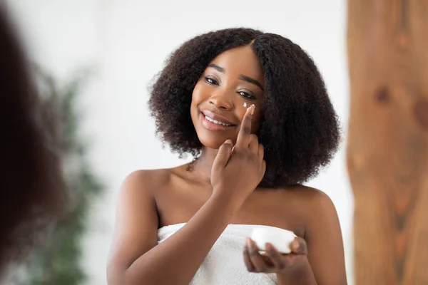 Atractiva joven Afro mujer aplicando bajo crema para los ojos después del baño cerca del espejo en casa. Concepto de bienestar doméstico — Foto de Stock