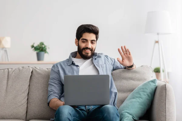 Online komunikace. Veselý arabský chlap mluví na notebook kamery, relaxaci na pohovce v domácím interiéru, volné místo — Stock fotografie