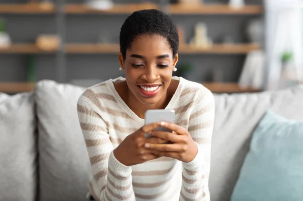 Bella app. Allegro giovane donna nera che si rilassa sul divano con smartphone — Foto Stock