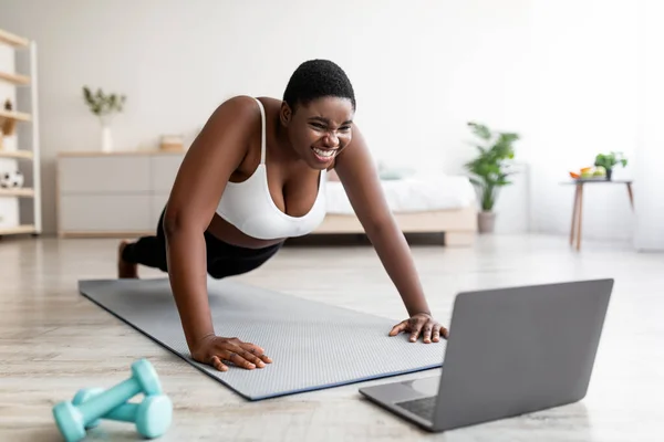 Plus size Afro woman standing in plank pose or doing push up near laptop, working out to online sports tutorial at home — стоковое фото