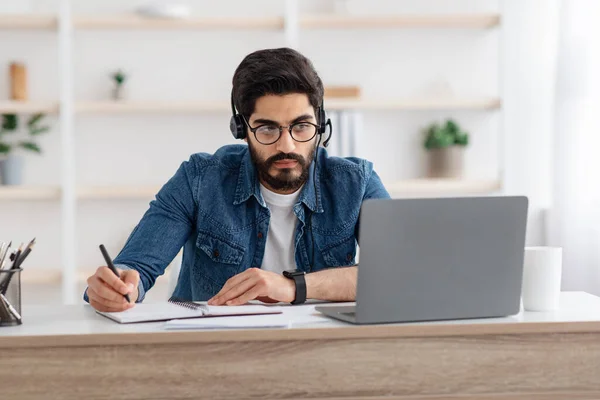 Millennial arab τύπος σε ακουστικά βλέποντας σε απευθείας σύνδεση webinar στο φορητό υπολογιστή στο σπίτι, μελετώντας με τον υπολογιστή και κρατώντας σημειώσεις — Φωτογραφία Αρχείου