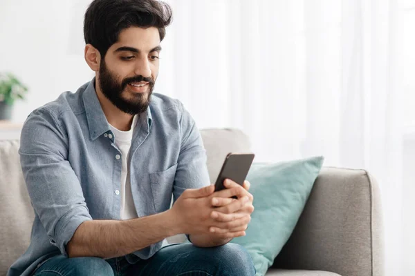 Comunicação móvel. Homem árabe feliz ler mensagens no smartphone em casa enquanto relaxa no sofá, espaço livre — Fotografia de Stock