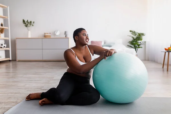 Señora negra curvilínea sentada cerca de la pelota de fitness, haciendo ejercicio en la alfombra deportiva en casa, longitud completa —  Fotos de Stock