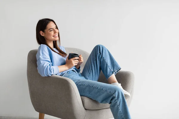 Retrato de mujer joven y feliz sentada en sillón con taza de café caliente, relajante sobre fondo blanco del estudio —  Fotos de Stock