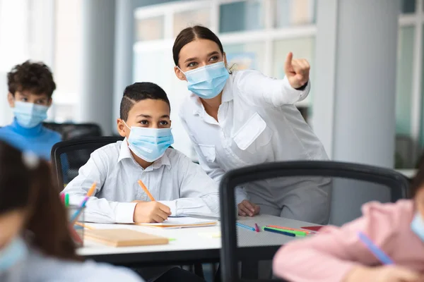 Profesora en mascarilla quirúrgica ayudando a un pequeño colegial hispano —  Fotos de Stock