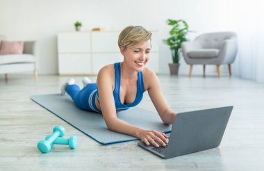 Gülümseyen kadın yoga minderinde dizüstü bilgisayar kullanarak yatıyor.
