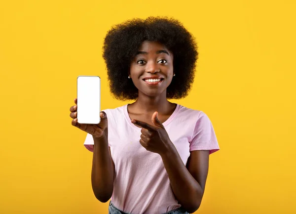 Cheerful african american lady pointing on smartphone with white empty screen in hand, standing over yellow background — Stock Photo, Image