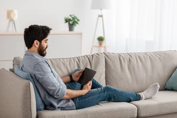 젊은 아라파 청년 이 디지탈 태블릿을 들고 거실 소파에 누워 복사 공간을 갖고 있습니다. — 스톡 사진