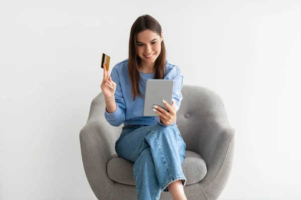 Διαδικτυακά ψώνια. Χαρούμενη κυρία με tablet και πιστωτική κάρτα παραγγελία πράγματα σε απευθείας σύνδεση, κάθεται στην πολυθρόνα σε λευκό φόντο — Φωτογραφία Αρχείου