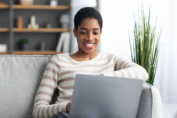 Lavoro a distanza. Sorridente signora freelancer nero utilizzando il computer portatile mentre seduto sul divano — Foto Stock