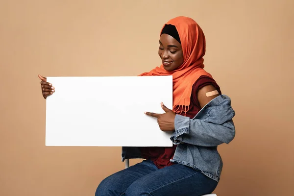 Mulher muçulmana negra positiva foi vacinada, apontando para o cartaz em branco — Fotografia de Stock