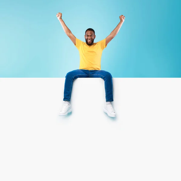 Plantilla publicitaria. Joven negro sentado encima de un afiche blanco en blanco con maqueta, levantando las manos —  Fotos de Stock