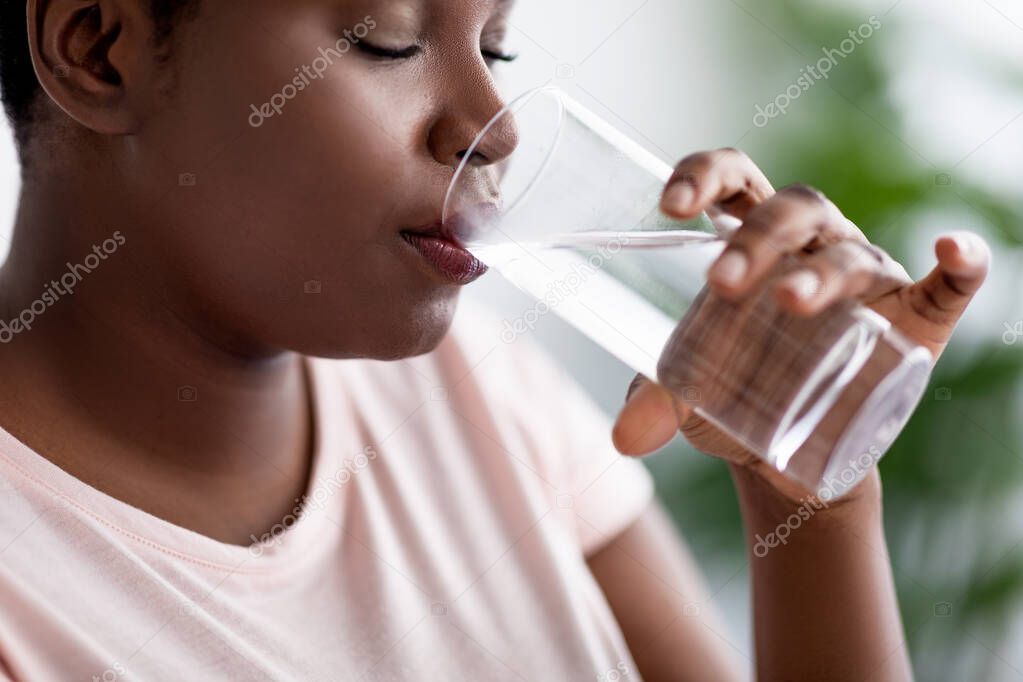 Keep hydrated for healthier living. Overweight black woman drinking clear mineral water from glass indoors