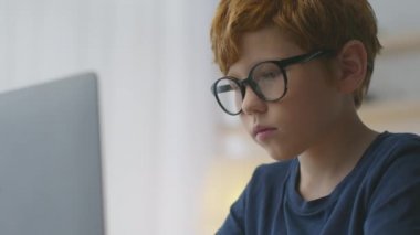 Bilgisayarda daktilo eden, evinden uzakta internetten ders çalışan odaklı kızıl saçlı çocuğun portresini kapat.
