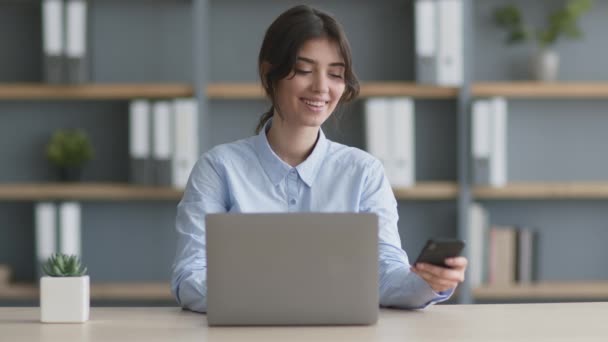 Junge Managerin tippt im Büro auf Laptop, bekommt und liest Nachricht auf Handy von geliebter Person — Stockvideo