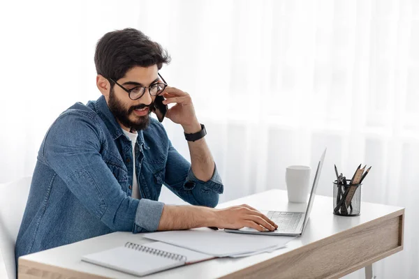 Trabajo desde casa. Árabe freelancer chico hablando por teléfono celular mientras trabaja con papeles y portátil, sentado en casa oficina — Foto de Stock