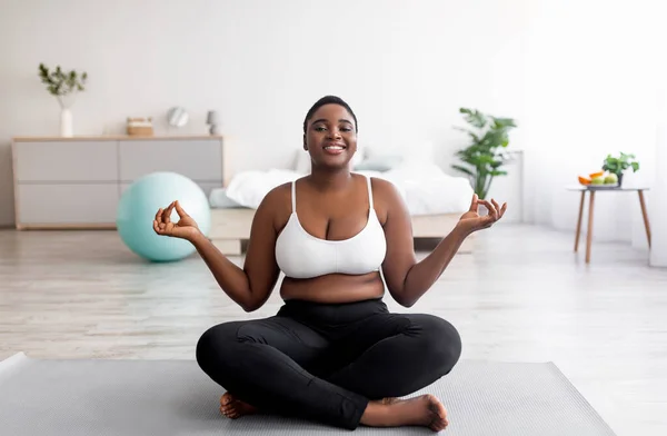 Mujer negra con sobrepeso sentada en alfombra deportiva en pose de loto, meditando, haciendo práctica de yoga en casa —  Fotos de Stock